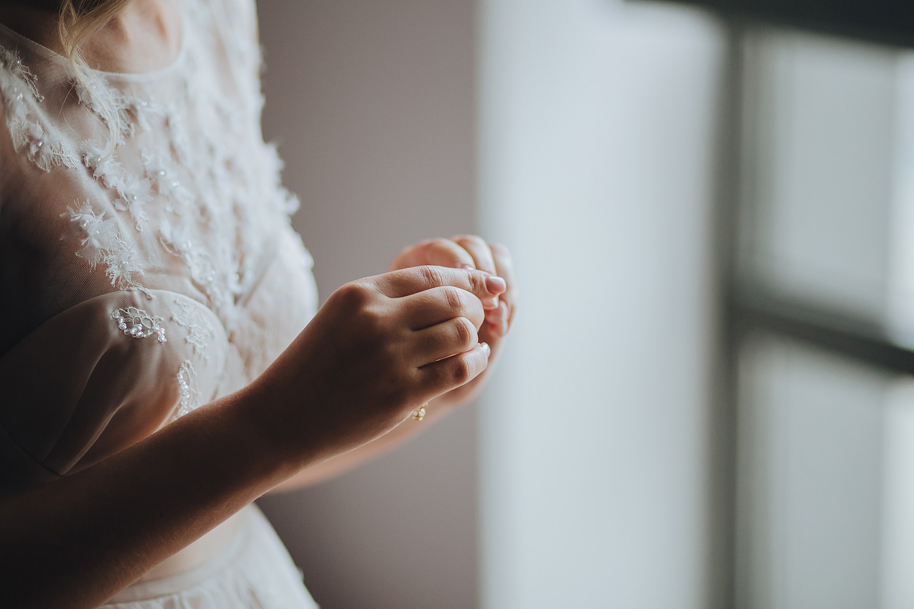 Ivy_1820_3 A woman in a white lace dress stands by a window, gently holding her hands together, with soft natural light illuminating her delicate dress and fingers.