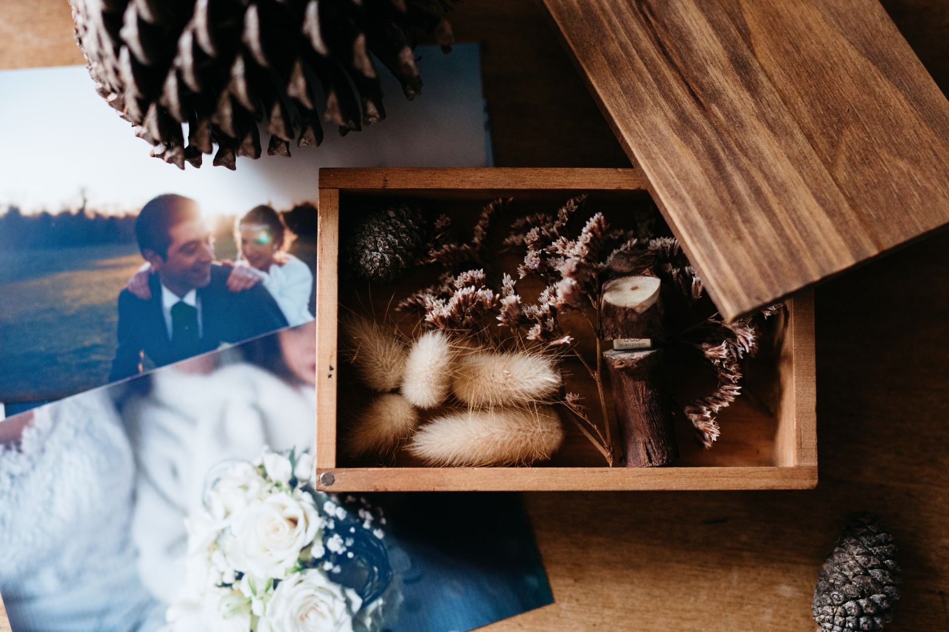 A wooden box with dried flowers and small pinecones sits open on a table, surrounded by printed photographs, including a wedding couple and a bouquet of white roses.