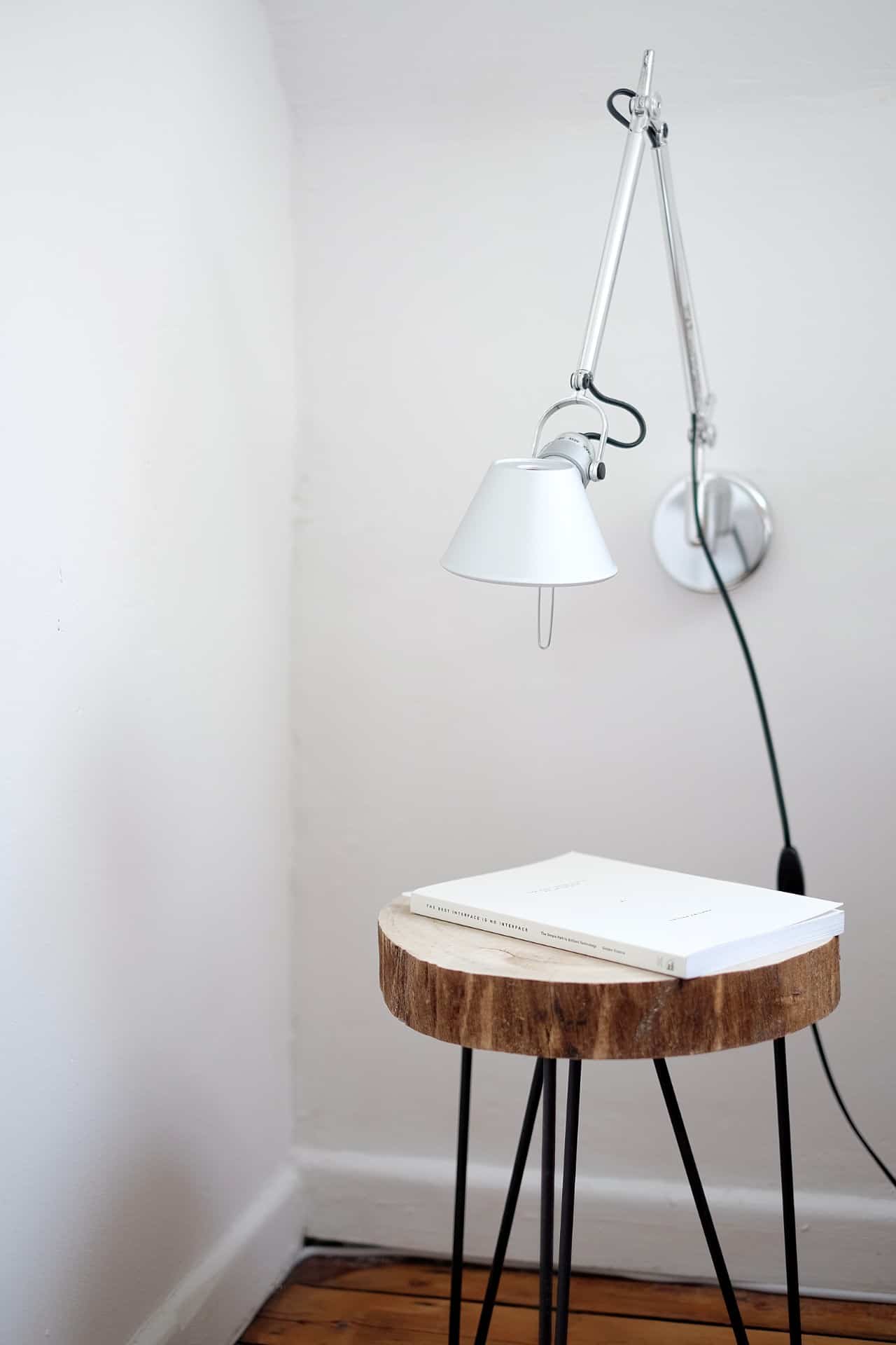 A minimalist corner with a white wall lamp, a small round wooden table with black metal legs, and two closed white books on top. The scene features simple, modern decor.
