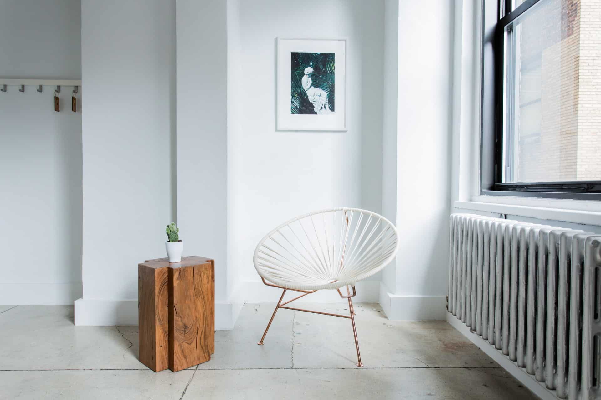 Minimalist room with a white modern chair, a small wooden table holding a potted plant, a radiator, and a framed artwork on the white wall near a large window.