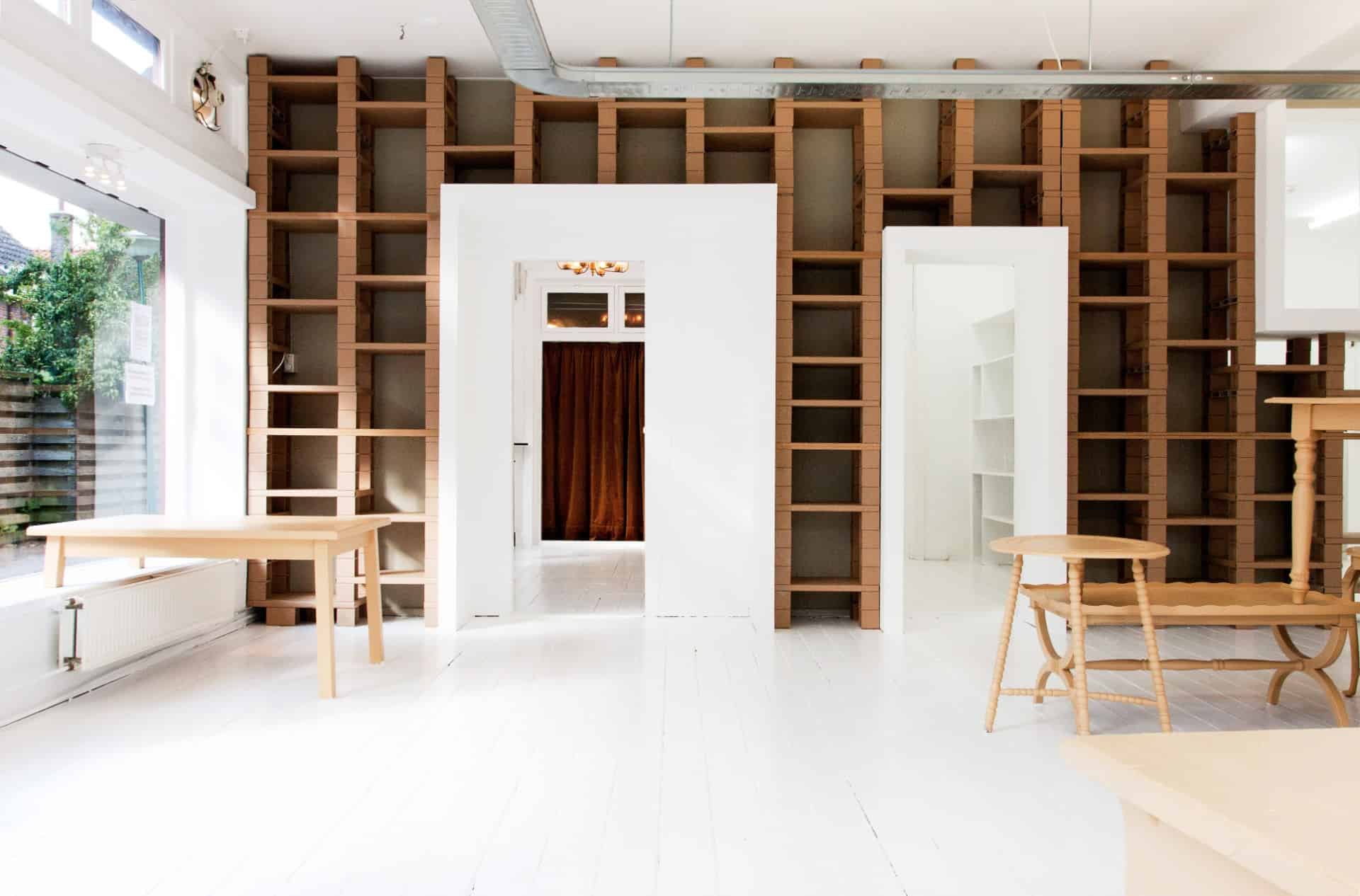 The_Design_Space_Interiors_71 Minimalist room with white floors, wooden shelves against the wall, a wooden table, chairs, and two doorways. Large windows on the left let in natural light, creating a bright, airy atmosphere.