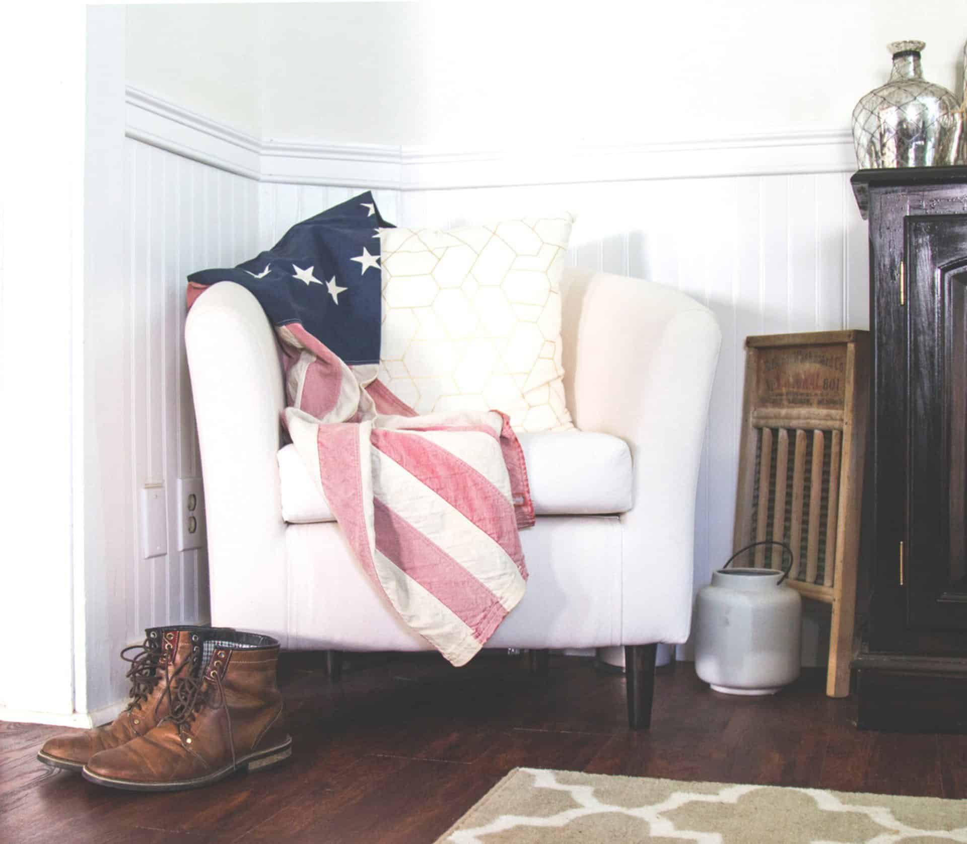 The_Design_Space_Interiors_75 A white armchair with an American flag blanket and pillows, brown boots on the floor, a small wooden crate, a jug, and a dark cabinet beside it, all on a hardwood floor.