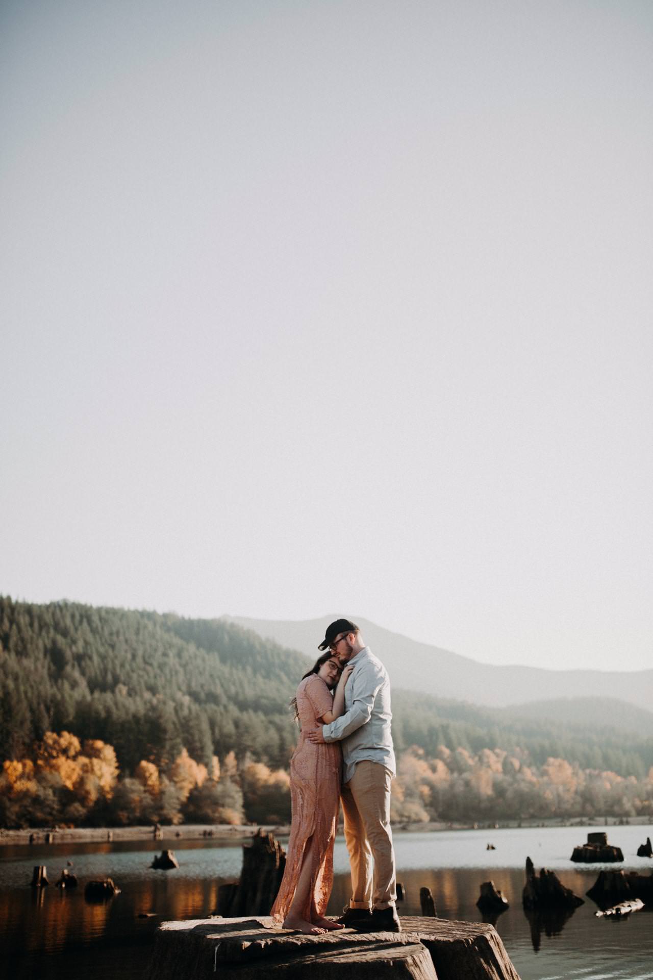A couple embraces on a large tree stump by a lake, surrounded by autumn trees and distant mountains under a clear sky.