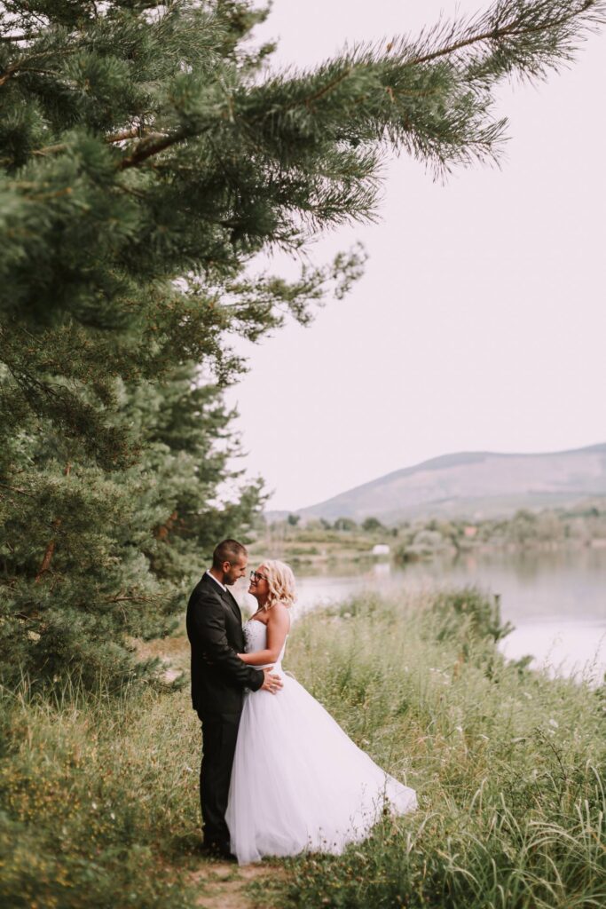 Bride and groom stand together outdoors by a lake, surrounded by tall grass and pine trees, with mountains visible in the background.