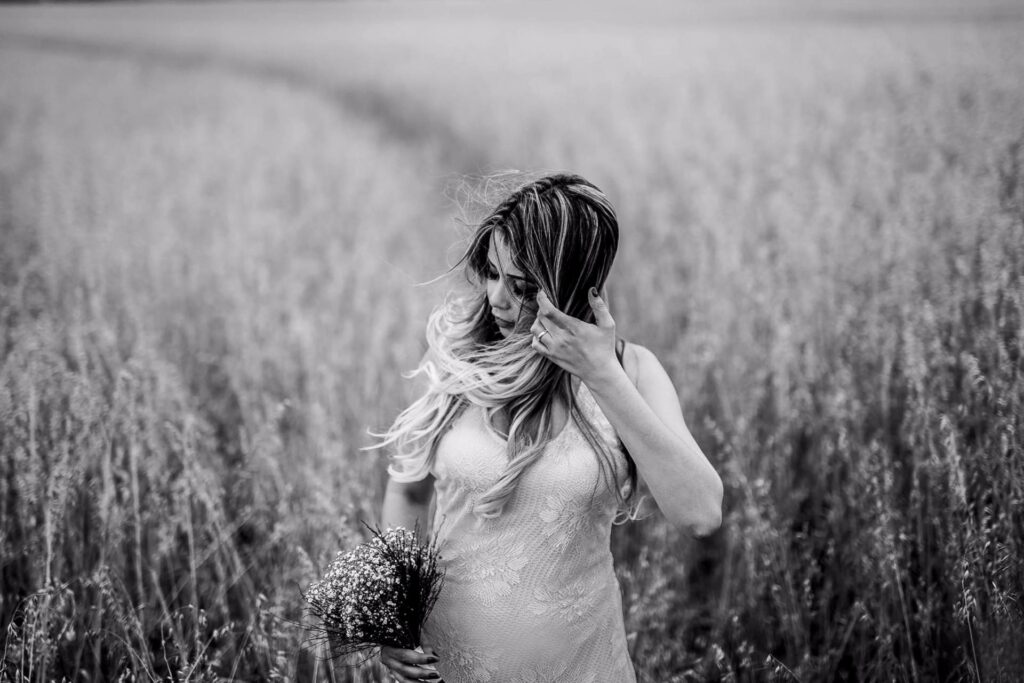 A woman in a light dress stands in a field of tall grass, holding a small bouquet of flowers and brushing her hair aside. The image is black and white.