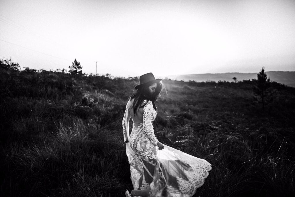 A woman in a lacy dress and hat stands in a grassy field at sunset, turning slightly as she looks back over her shoulder. The scene is in black and white, creating a dramatic and nostalgic mood.