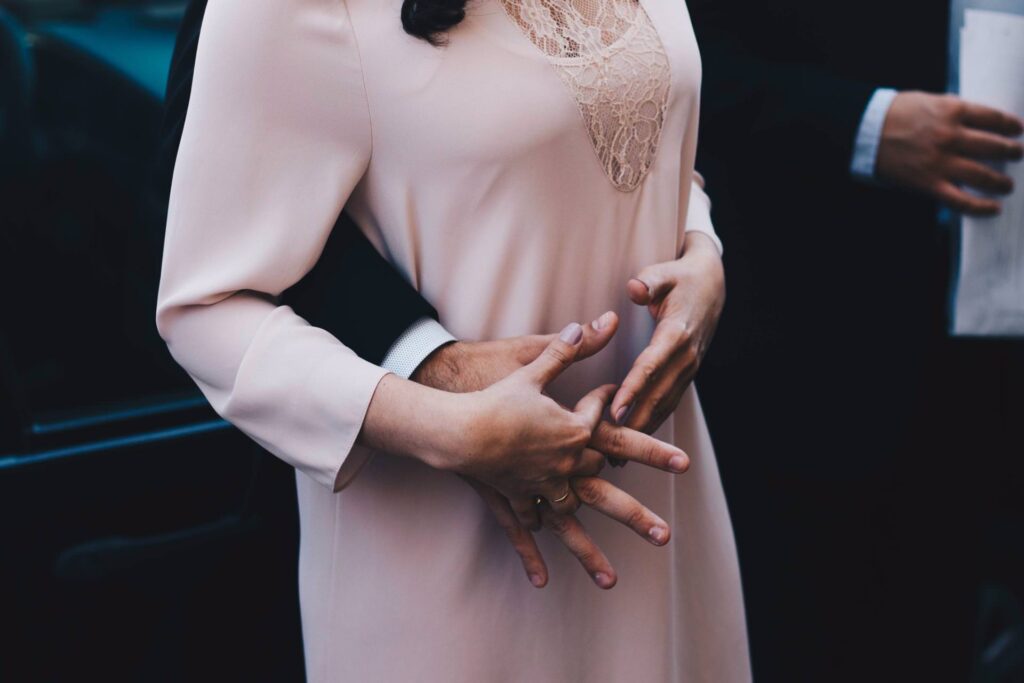 A person in a pale pink dress is gently embraced from behind by someone in a suit, their hands intertwined at the front. Another hand holding a paper appears on the right side of the image.