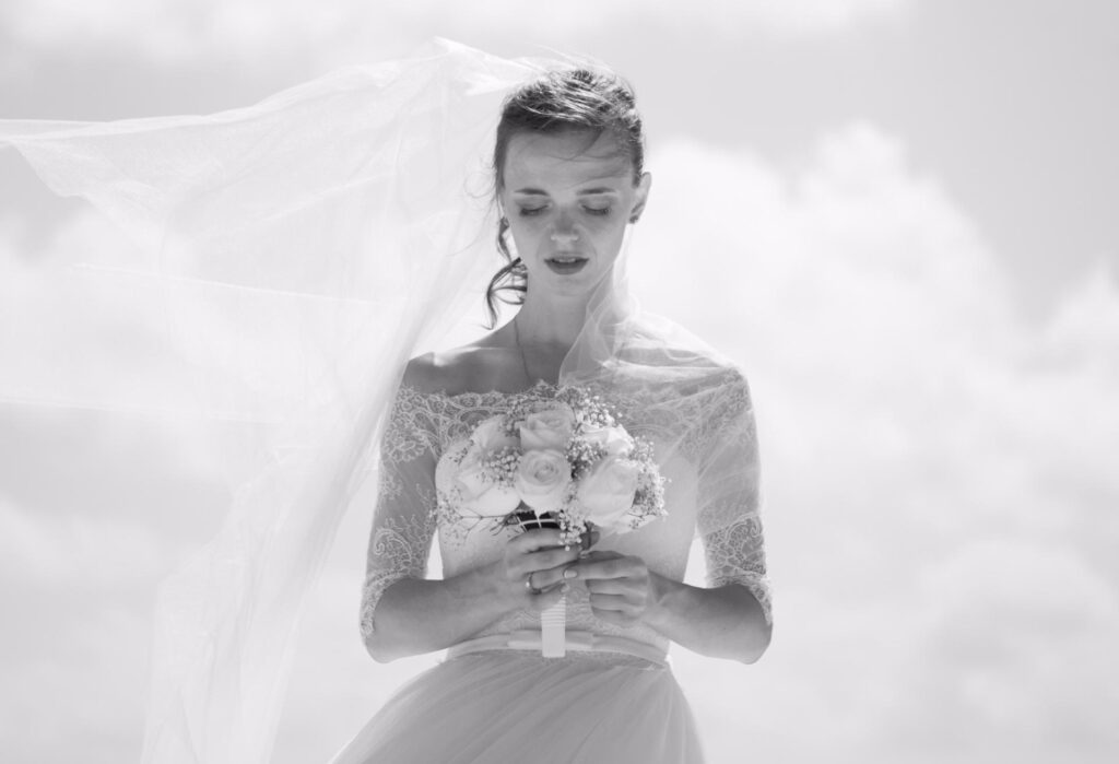 A bride in a lace wedding dress holds a bouquet of flowers and looks down, with her veil flowing in the wind against a bright, cloudy sky.