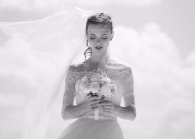 A bride in a lace wedding dress holds a bouquet of flowers and looks down, with her veil flowing in the wind against a bright, cloudy sky.