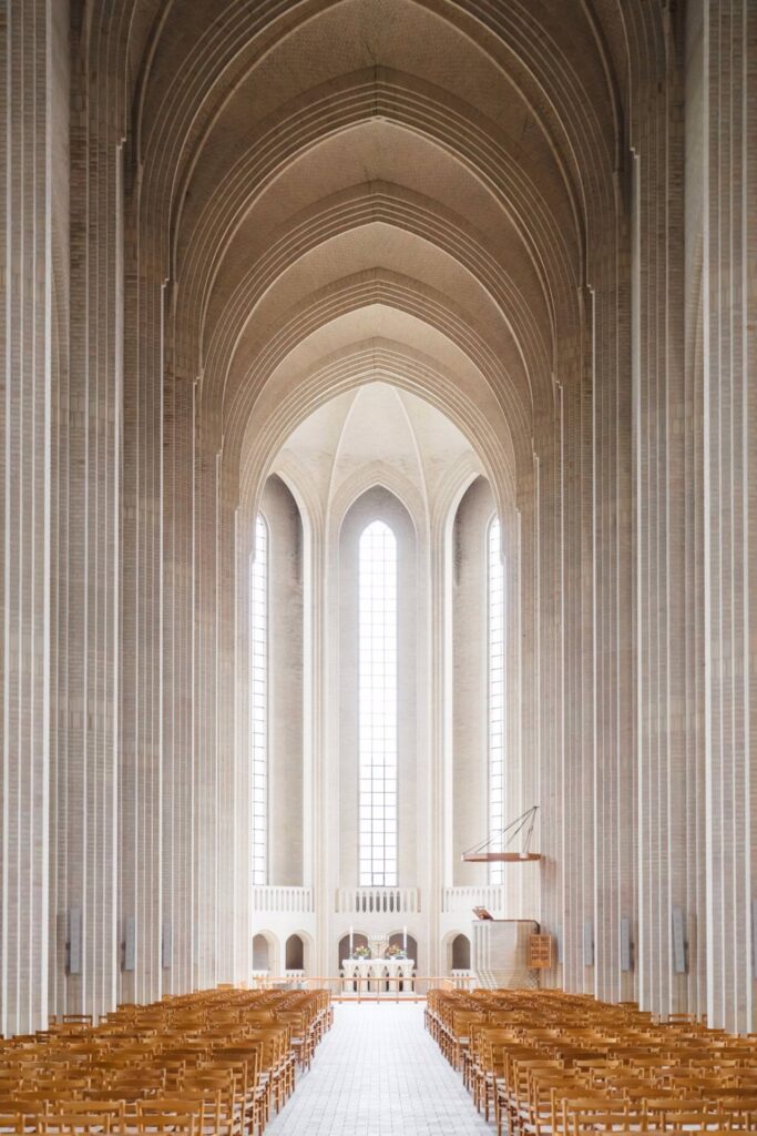 Tall, bright church interior with soaring arched ceilings, long rows of wooden chairs, tall windows letting in natural light, and an altar at the far end. The architecture is minimalist and elegant.