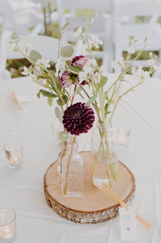 Glass bottles with purple and white flowers sit on a wooden slice centerpiece, surrounded by small votive candles and a Love tag, on a white tablecloth. White chairs are visible in the background.