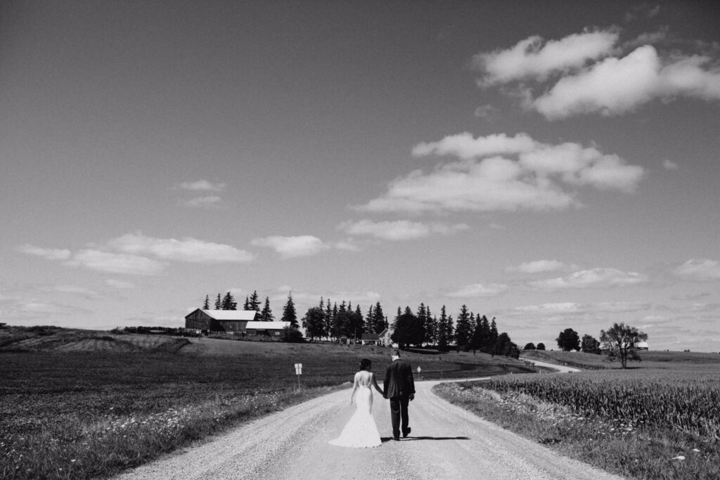 A bride and groom walk hand in hand down a rural gravel road toward a farmhouse, surrounded by fields and trees, under a partly cloudy sky.