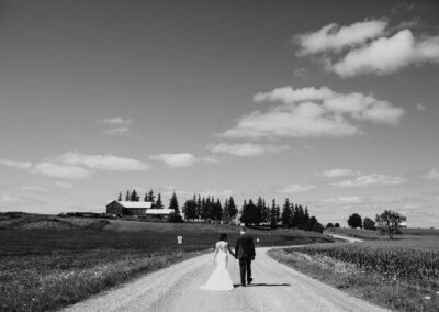 A bride and groom walk hand in hand down a rural gravel road toward a farmhouse, surrounded by fields and trees, under a partly cloudy sky.