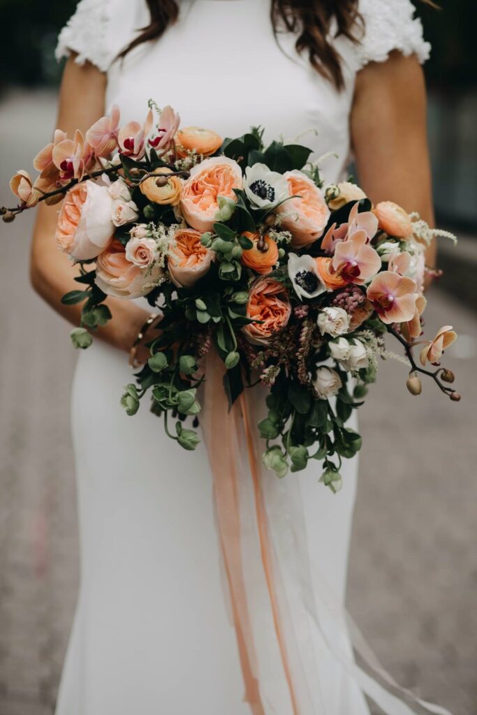 A bride in a white dress holds a large bouquet with peach roses, white anemones, pink orchids, and greenery, tied with long ribbons.