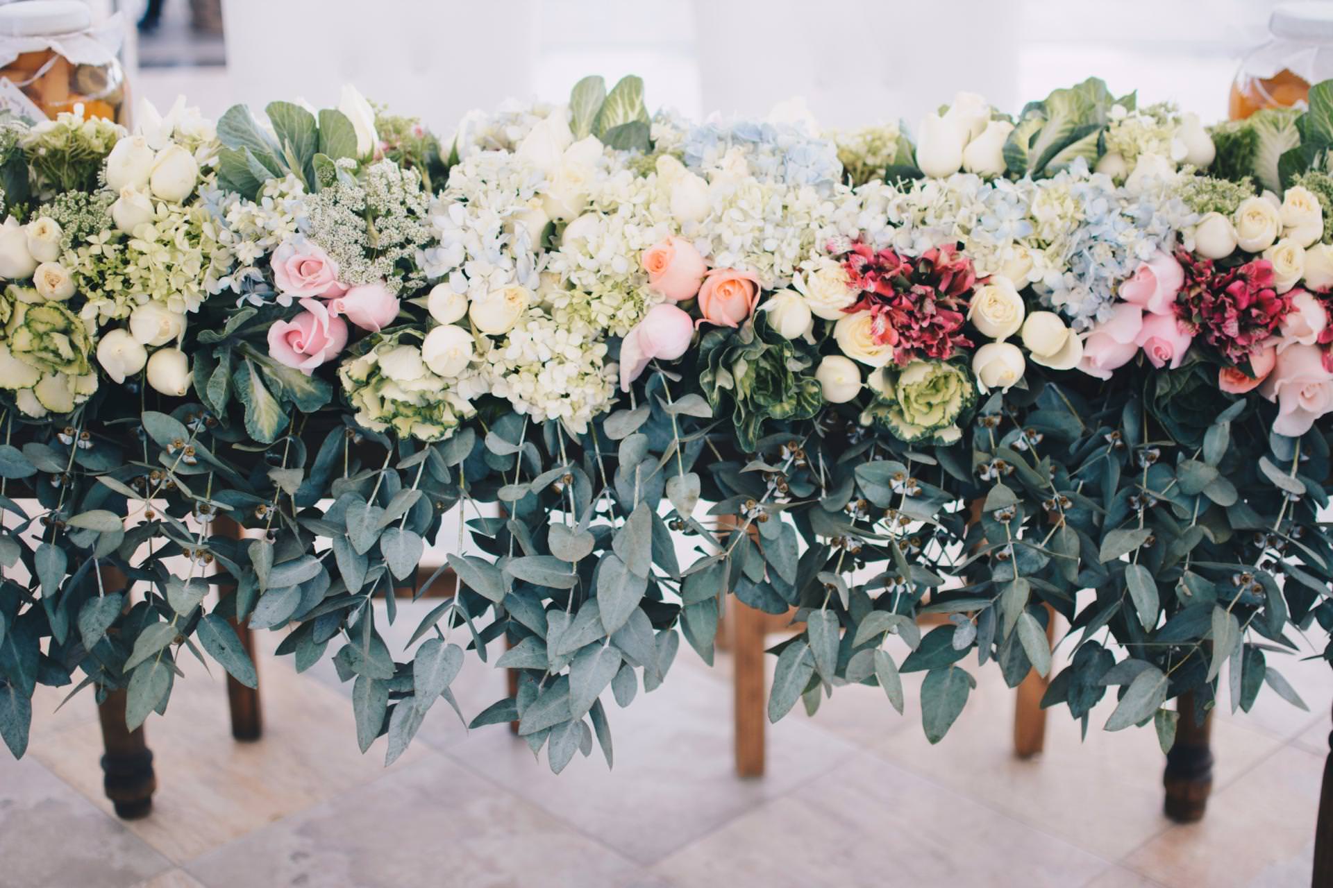 A lush flower arrangement with white, pink, and peach roses, hydrangeas, and greenery drapes over the edge of a table, creating an elegant, garden-inspired centerpiece.