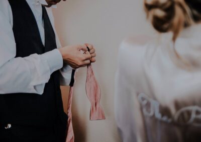 A person in formalwear adjusts a pink bow tie, while another person in a light robe with cursive writing on the back is visible in the foreground, facing away.