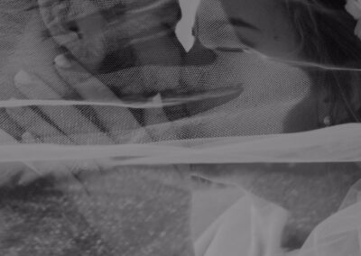 A bride and groom share an intimate moment, faces close together under a sheer wedding veil, smiling softly at each other.