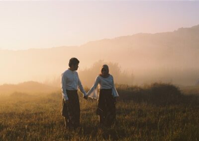 Two people wearing traditional clothing hold hands while walking through a grassy field at sunrise, with mist and distant hills in the background.