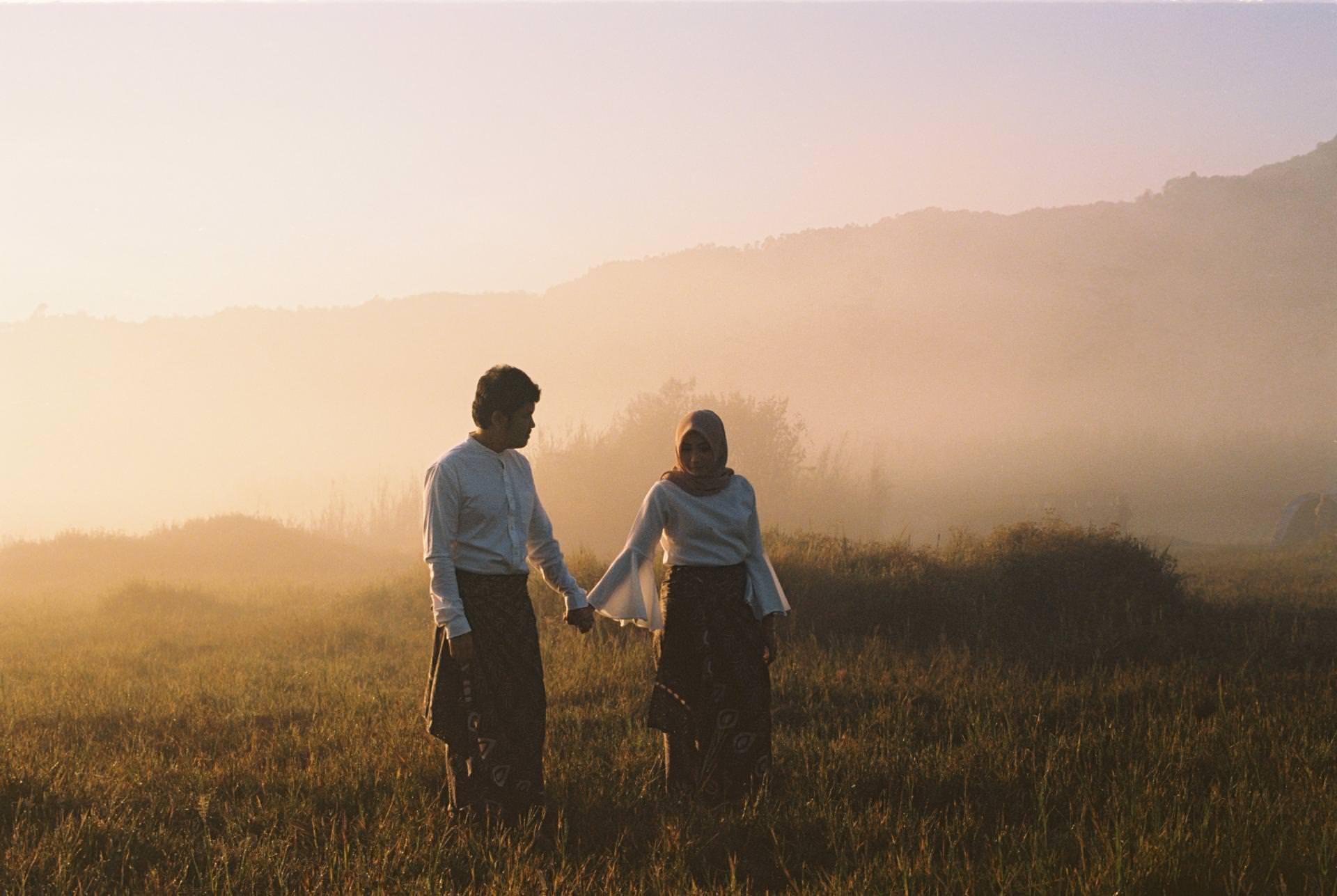 Two people wearing traditional clothing hold hands while walking through a grassy field at sunrise, with mist and distant hills in the background.