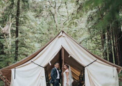 A bride and groom stand inside a canvas tent in a forest, surrounded by tall trees. The bride holds a bouquet and wears a veil, while the groom is in a suit. The scene is serene and rustic.