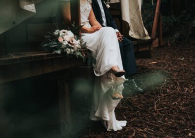 A bride and groom sit close together on a wooden bench outdoors. The bride holds a bouquet of flowers, and her dress and shoes are visible. The groom wears a dark suit. The ground is covered in brown leaves.