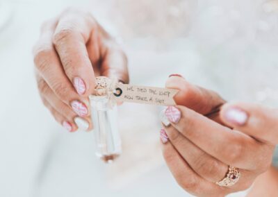 A person with pink patterned nails holds a small glass bottle with a cork and a tag that reads, We tied the knot, now take a shot..