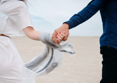Two people, one in a white dress and the other in a blue shirt, hold hands on a sandy beach. The image is cropped to show only their torsos and clasped hands, suggesting connection and togetherness.