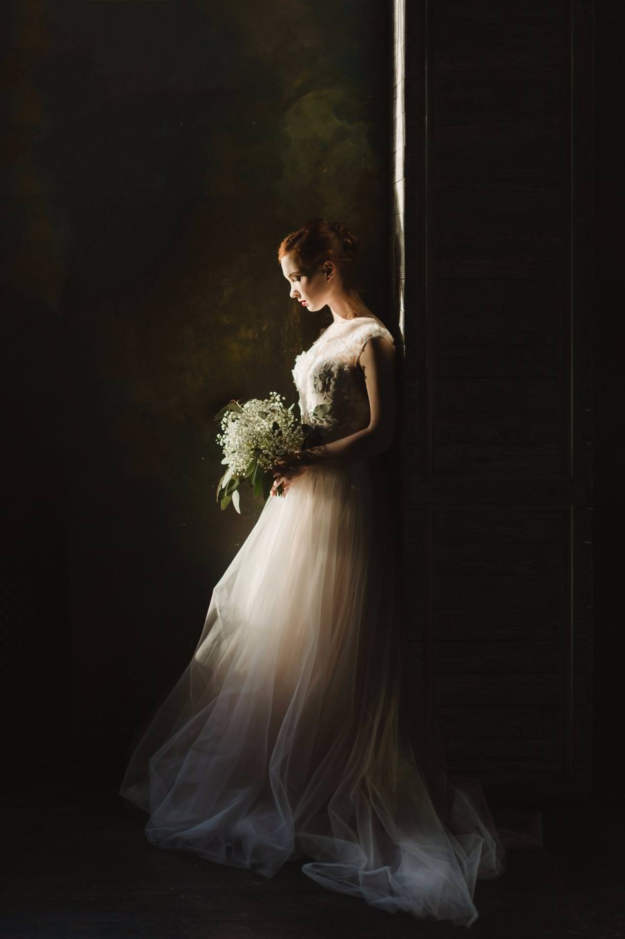 A woman in a flowing white wedding dress stands in soft light by a dark wall, holding a bouquet of white flowers and looking down thoughtfully.