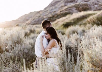 A couple embraces in a sunlit field with tall grass and hills in the background, creating a warm, intimate atmosphere.