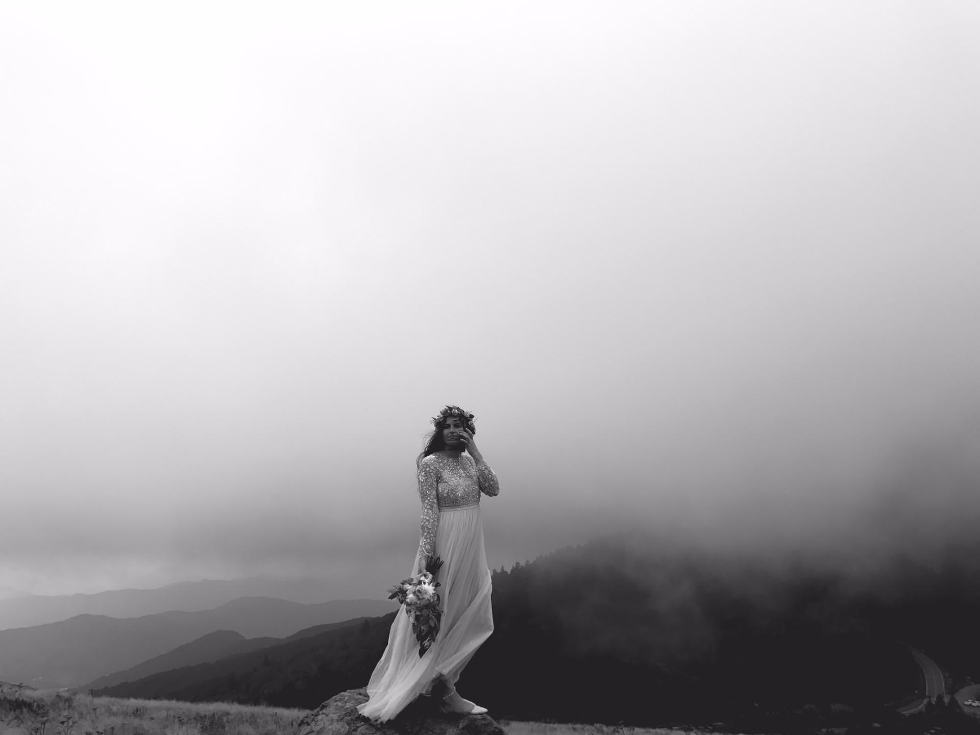 A woman in a long dress and floral crown holds a bouquet, standing on a rock with mountains and misty clouds in the background. The image is black and white.