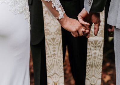 A bride and groom hold hands during a wedding ceremony, standing before an officiant wearing a decorative stole with gold embroidery.