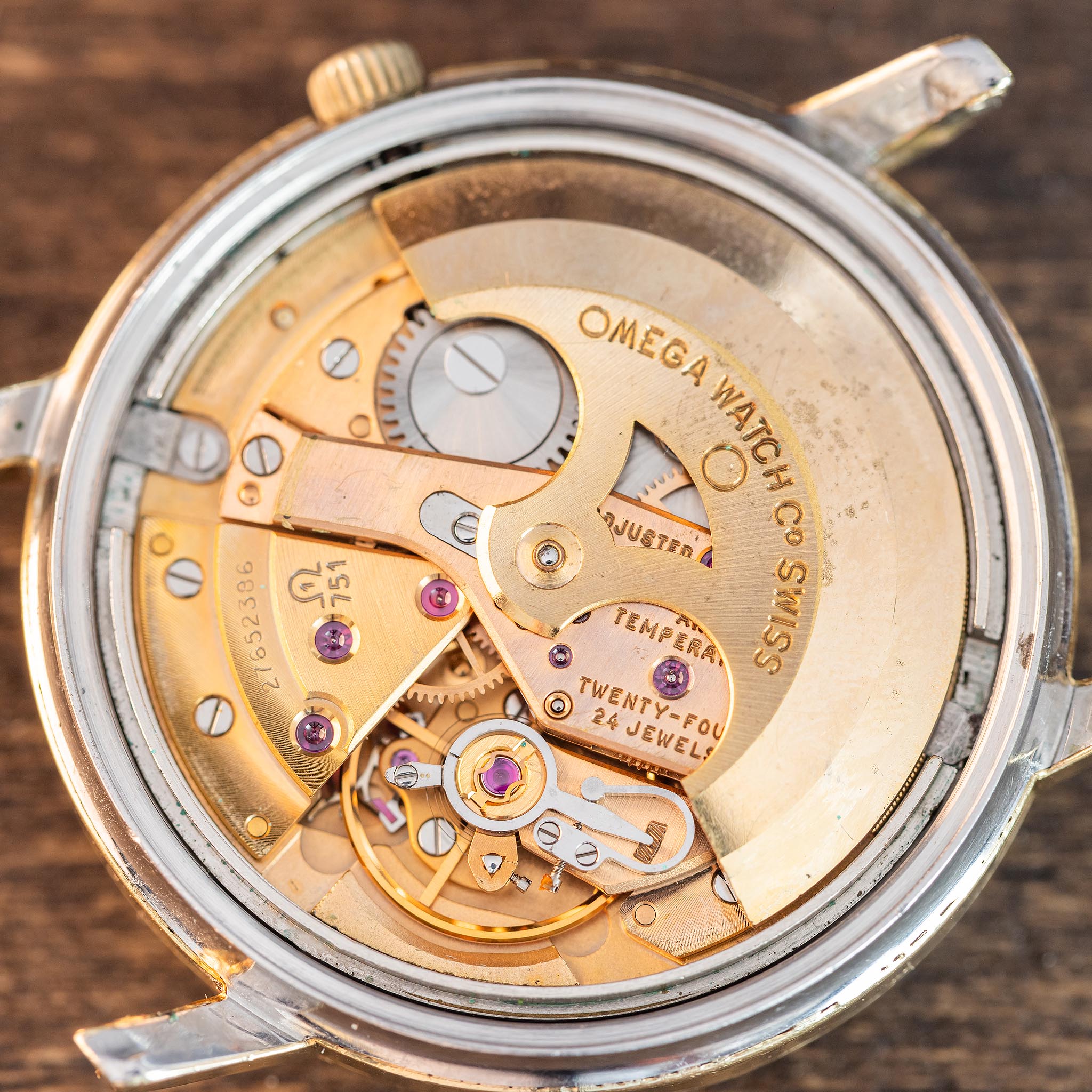 Close-up of the internal mechanism of an Omega wristwatch, showcasing gold-toned gears and intricate components from the renowned Omega caliber 751 movement. The engraved text OMEGA WATCH CO SWISS is clearly visible on the 24 jewel movement, all set against a wooden background.