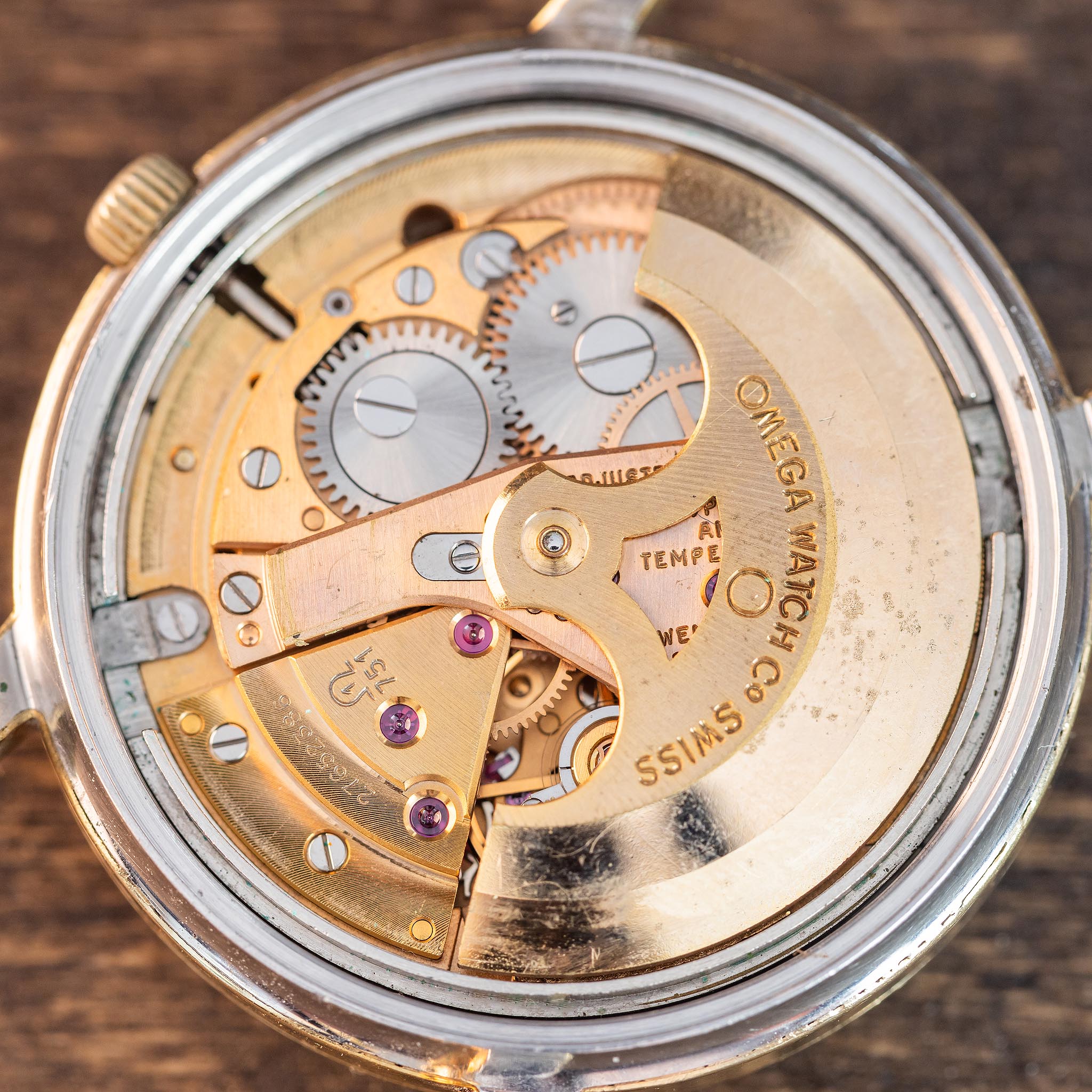 Close-up of the inner mechanical movement of an Omega wristwatch, showcasing gears, screws, and engraved text on gold-toned components from a vintage Omega automatic movement, set against a wooden surface.