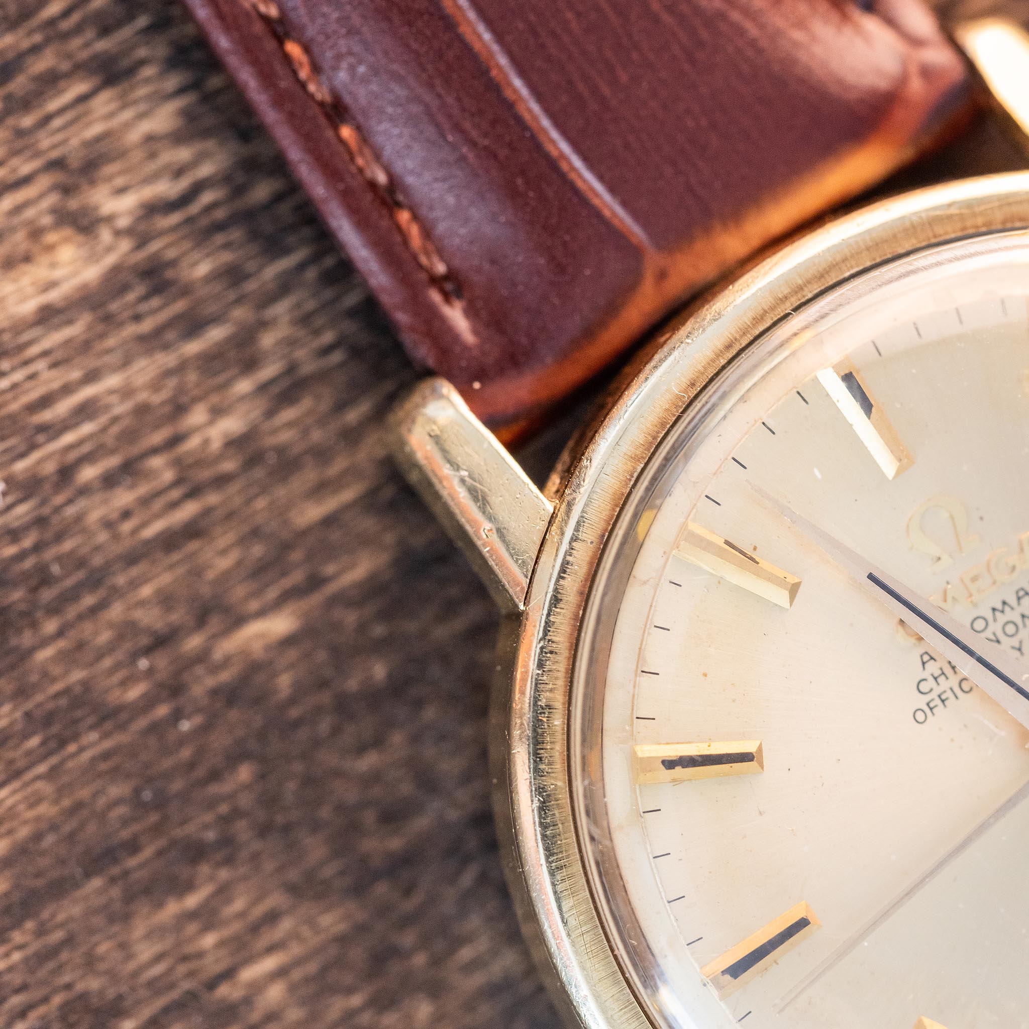 Close-up of a vintage Omega Constellation with a brown leather strap on a wooden surface, showing the gold case, part of the face, and hour markers—a classic example of a Swiss automatic watch.