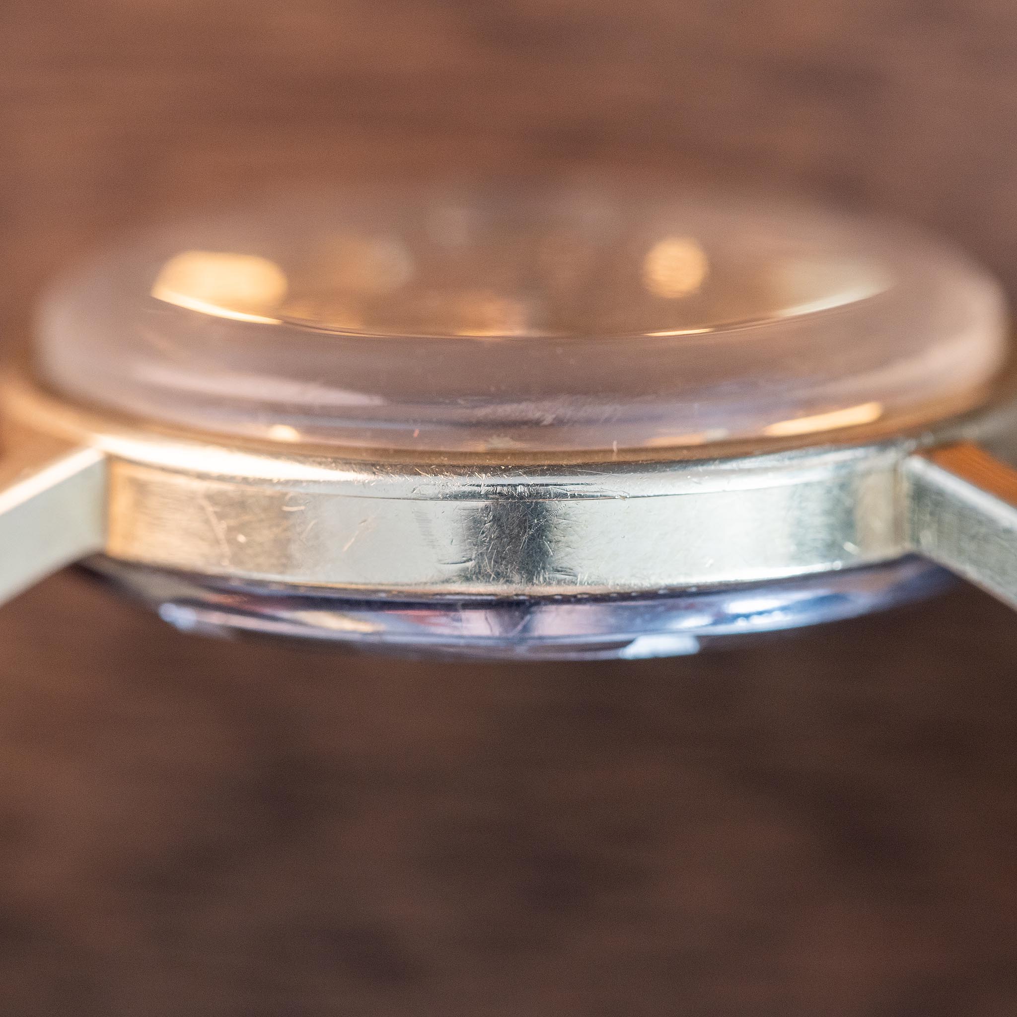 Close-up of the side of a gold-colored vintage Swiss watch case with a clear, domed crystal, showing some scratches and wear on the metal surface; background is blurred brown.