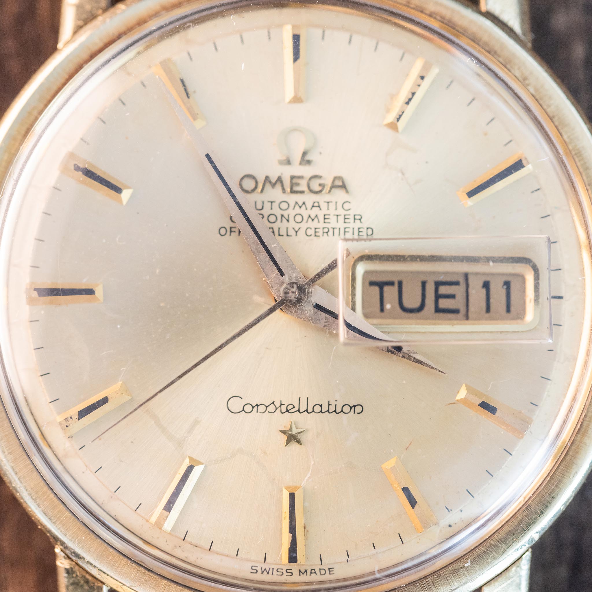 A close-up of an Omega Constellation watch face showing the time as 2:35:38 and the date as Tuesday the 11th. This vintage Swiss watch features gold hour markers and hands on a cream background, highlighting its timeless elegance.