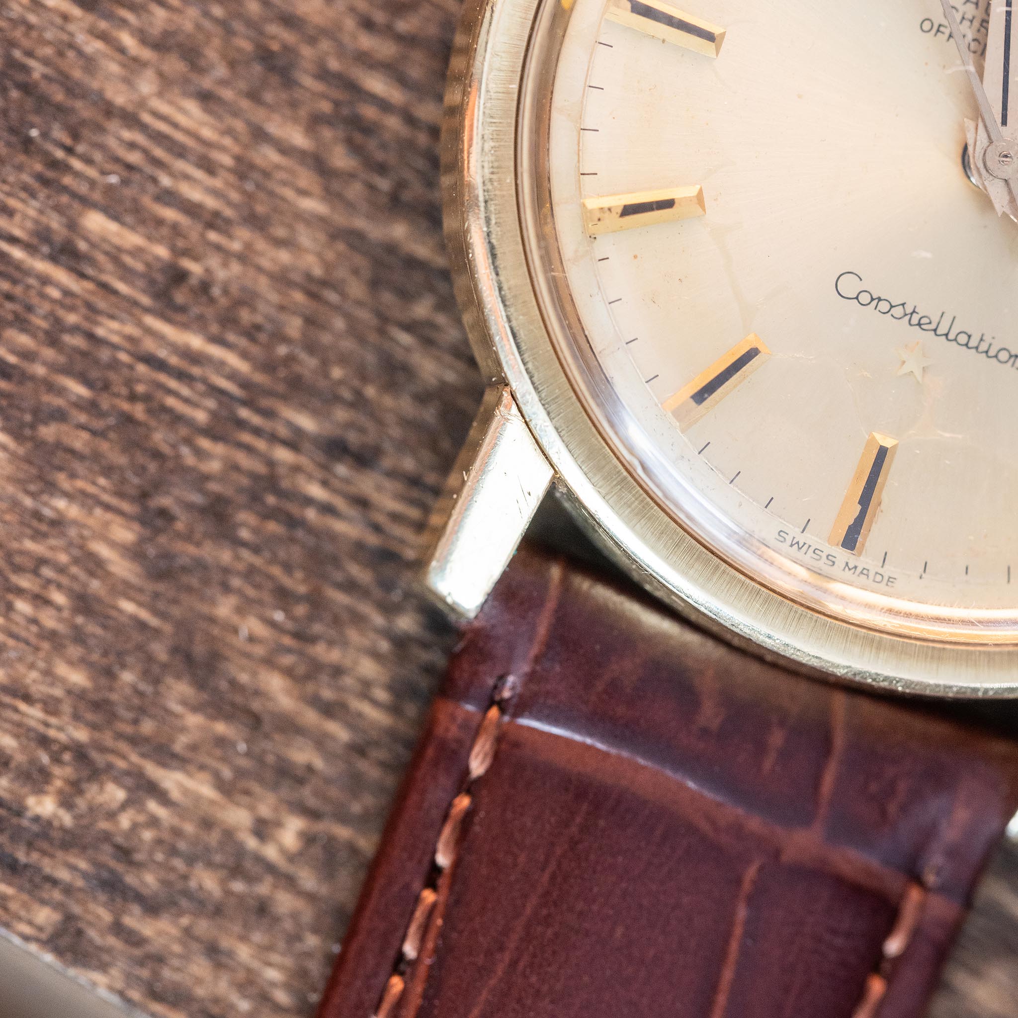 Close-up of a vintage Omega Constellation wristwatch with a gold bezel, cream dial, brown leather strap, and visible Constellation and Swiss Made text. The watch rests on a wooden surface.