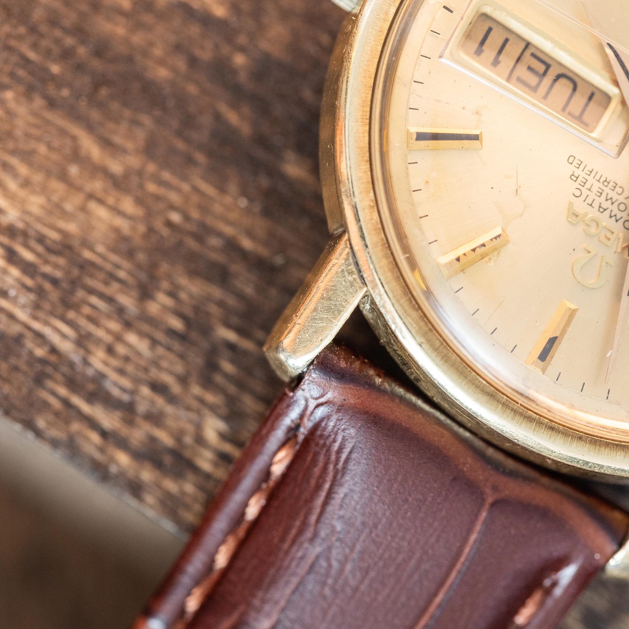 A close-up of a vintage Omega Constellation wristwatch with a gold case and brown leather strap, showing the day as TUE and part of the dial on a wooden surface.