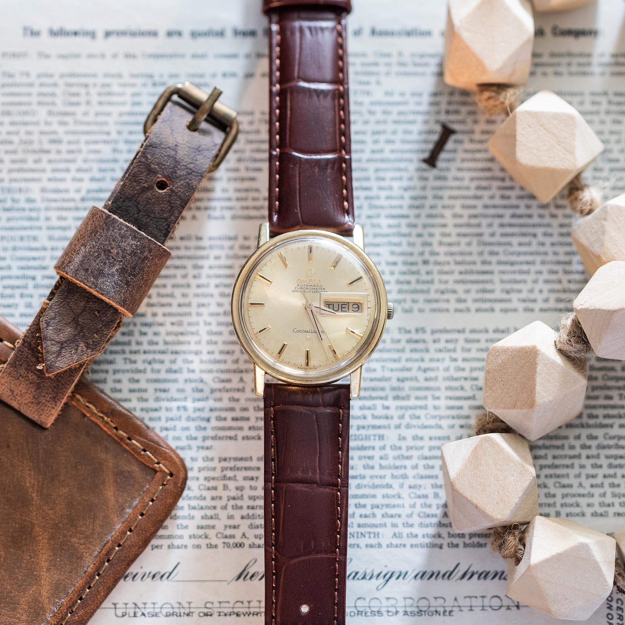 A vintage Omega Constellation gold wristwatch with a brown leather strap is placed on a newspaper beside an additional leather strap and a decorative string of wooden beads.