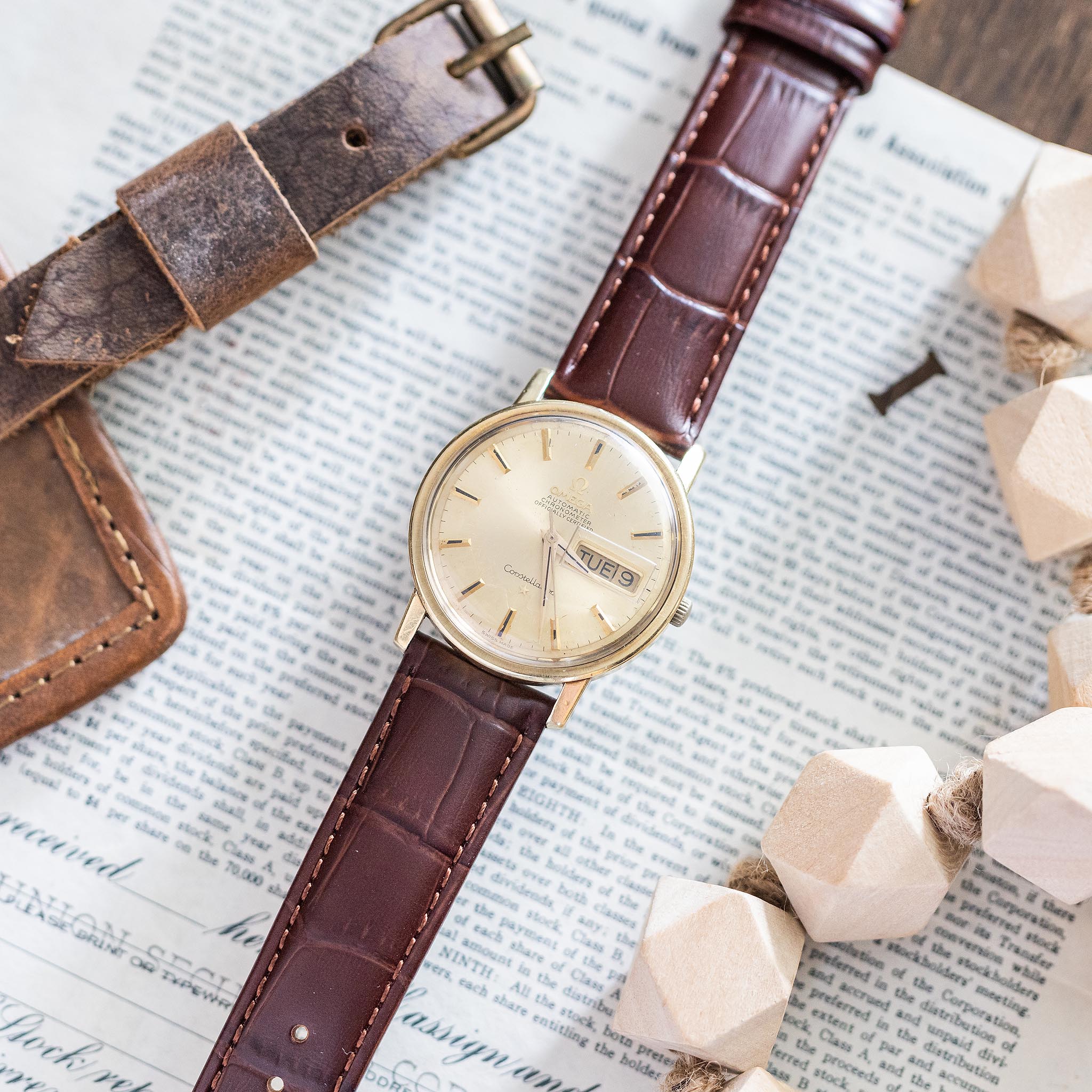A vintage Omega Constellation gold wristwatch with a brown leather strap rests on a document, surrounded by a matching leather strap and geometric wooden beads. This classic vintage Swiss watch displays both the day and date, adding timeless charm to its elegant design.