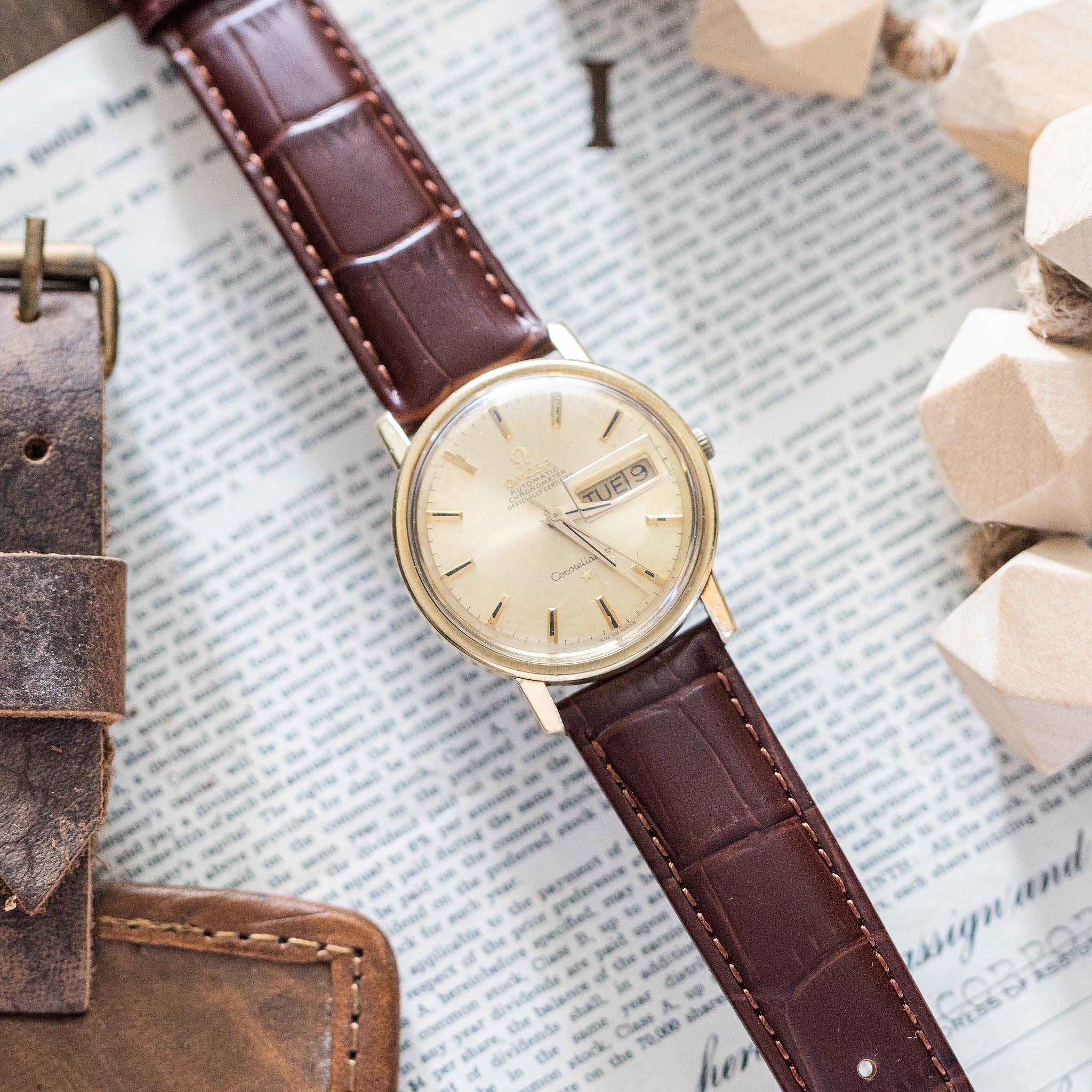 A vintage Omega Constellation with a gold case and brown leather strap rests elegantly on an open book, surrounded by a leather item and wooden beads. This distinguished Swiss automatic watch displays both the day and date on its face, highlighting its timeless sophistication.