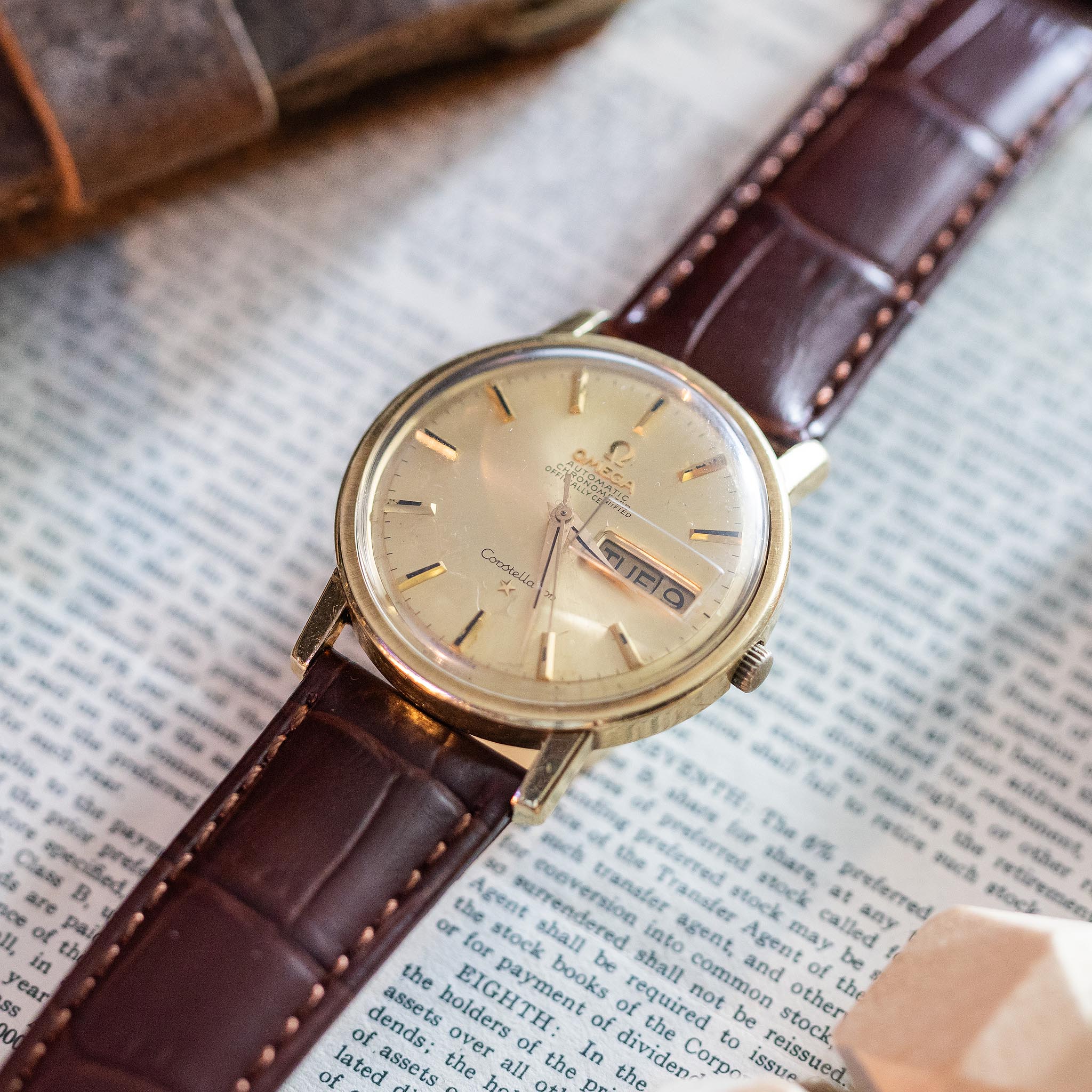 A vintage Omega Constellation wristwatch with a gold face and brown leather strap rests on an open book. This iconic vintage Swiss watch displays the date and day, complemented by elegant gold hour markers.