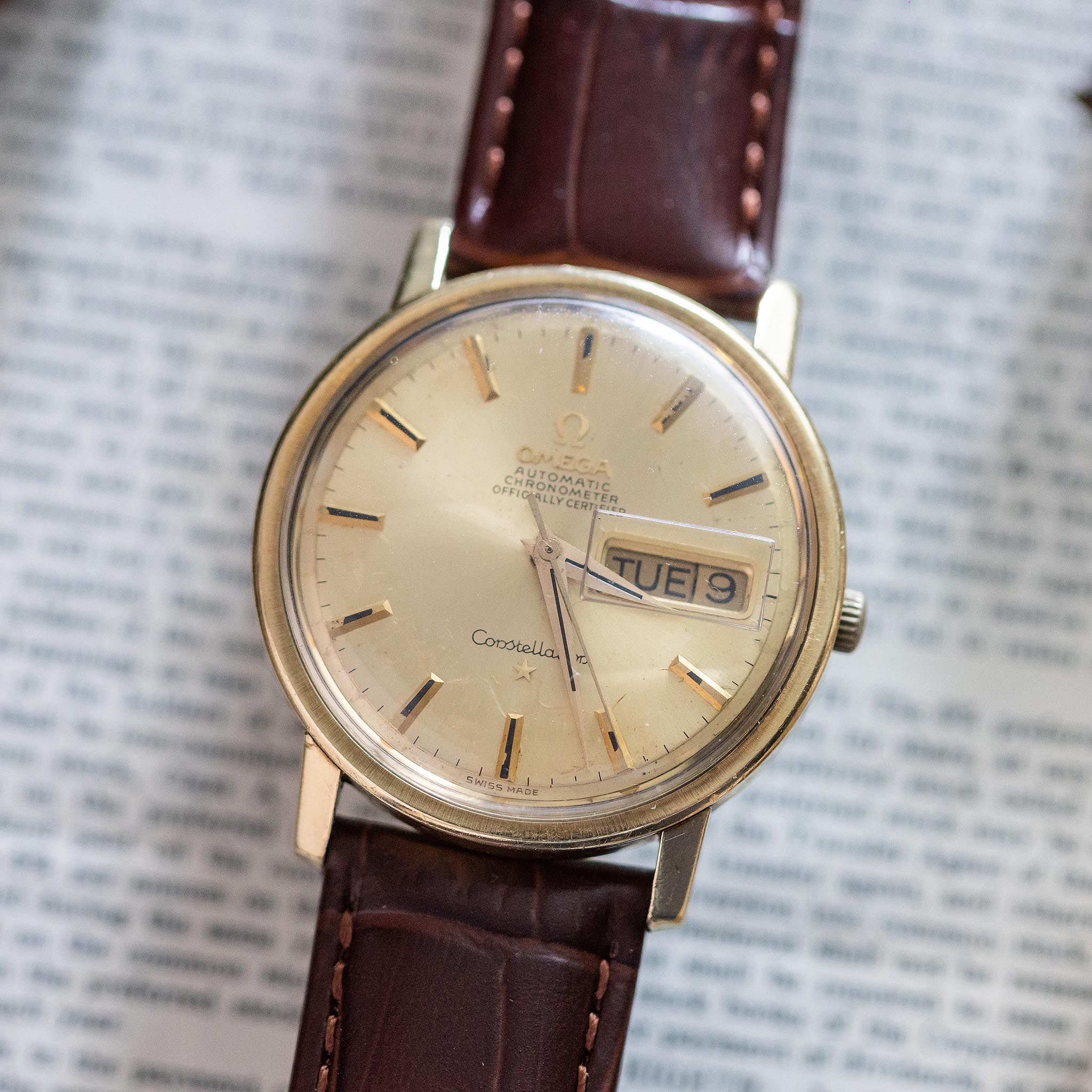 A vintage Omega watch with a gold case and brown leather strap sits on an open book. The classic watch face displays the day and date—showing TUE 9—and features gold hour markers and hands, highlighting the timeless elegance of this vintage Swiss watch.