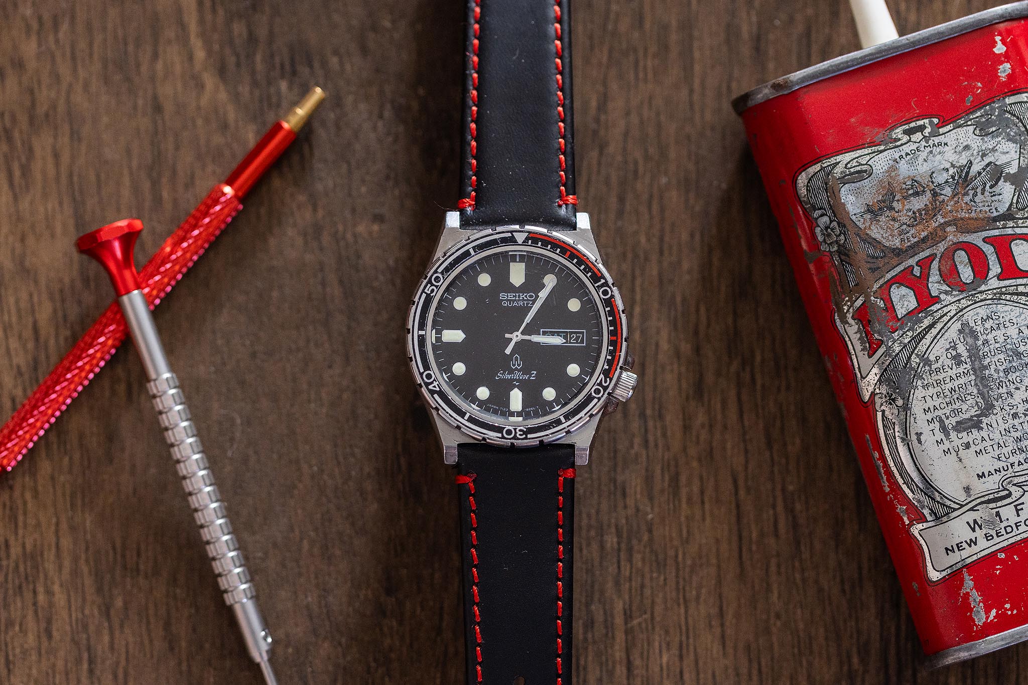 A vintage Seiko sports watch with a black dial and red-accented strap rests on a wooden surface, next to a red pen-like tool and a vintage red can with a white straw. A vintage Seiko sports watch with a black dial and red-accented strap rests on a wooden surface, next to a red pen-like tool and a vintage red can with a white straw.