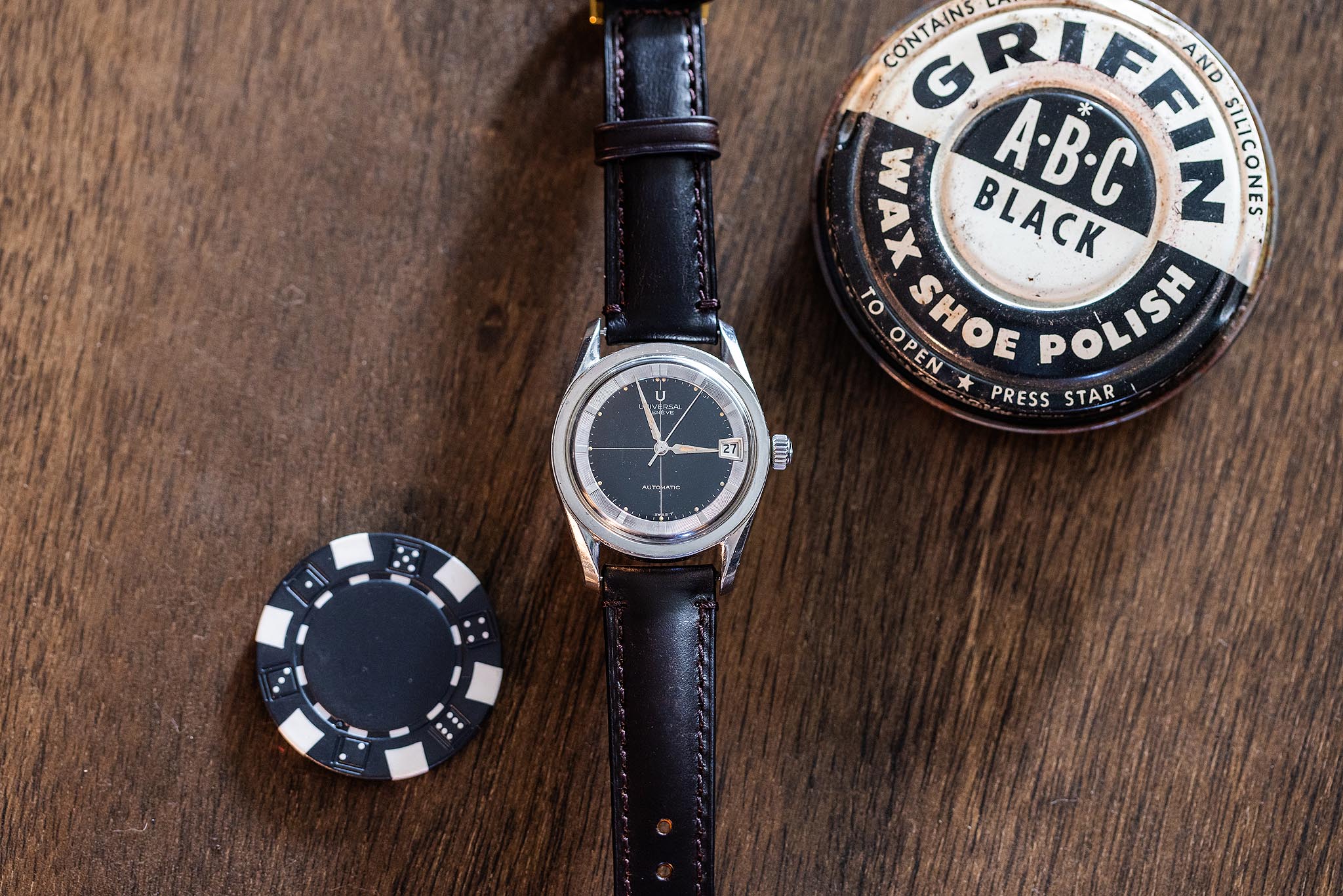 A Universal Genève Polerouter date 204612-2 wristwatch with a black leather strap rests on a wooden surface near a poker chip, a can of AX Shoe Polish, and a metal lighter.