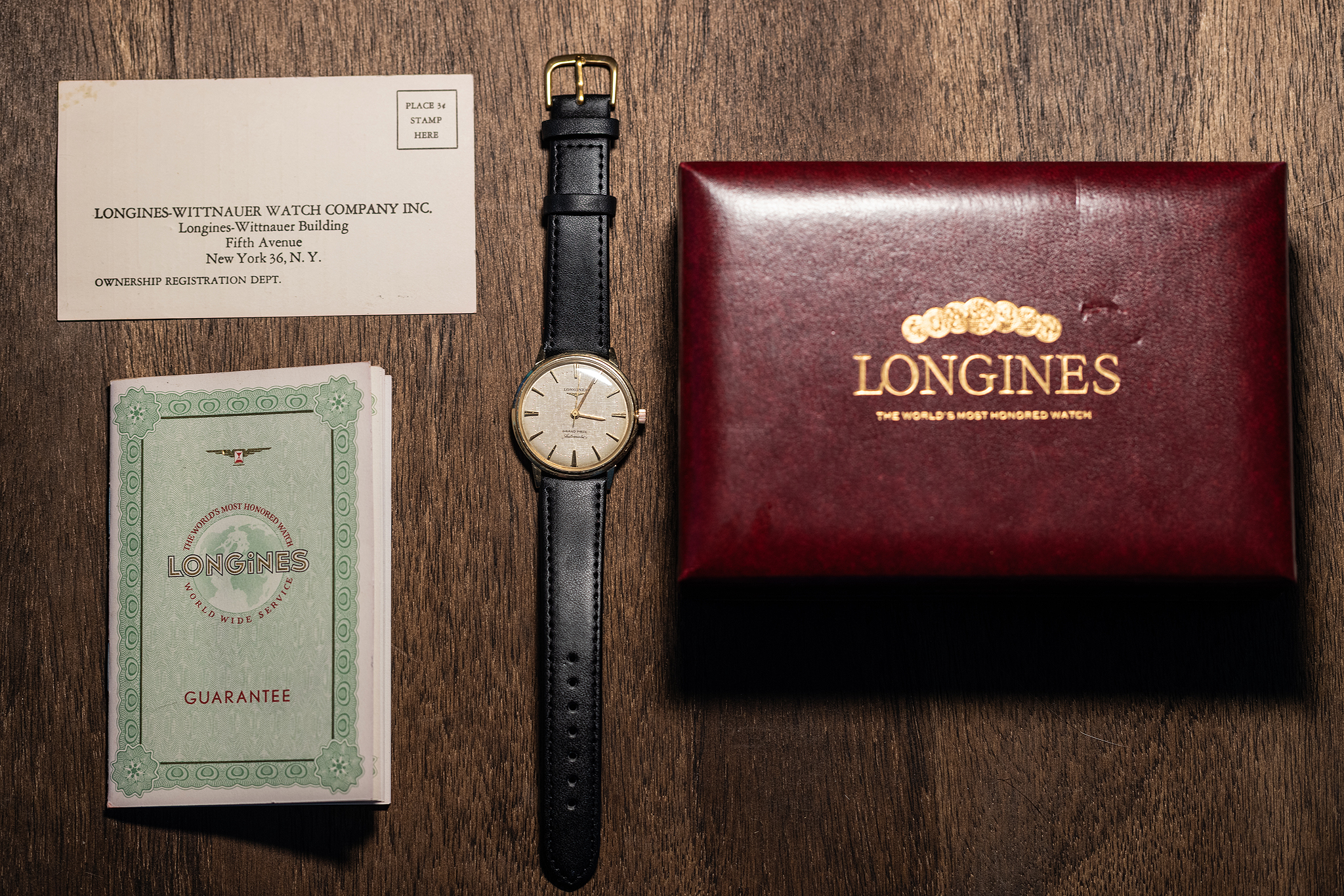 A vintage 14k gold Longines wristwatch with a black leather strap, positioned between its original guarantee booklet and a maroon presentation box, with an ownership registration card above.
