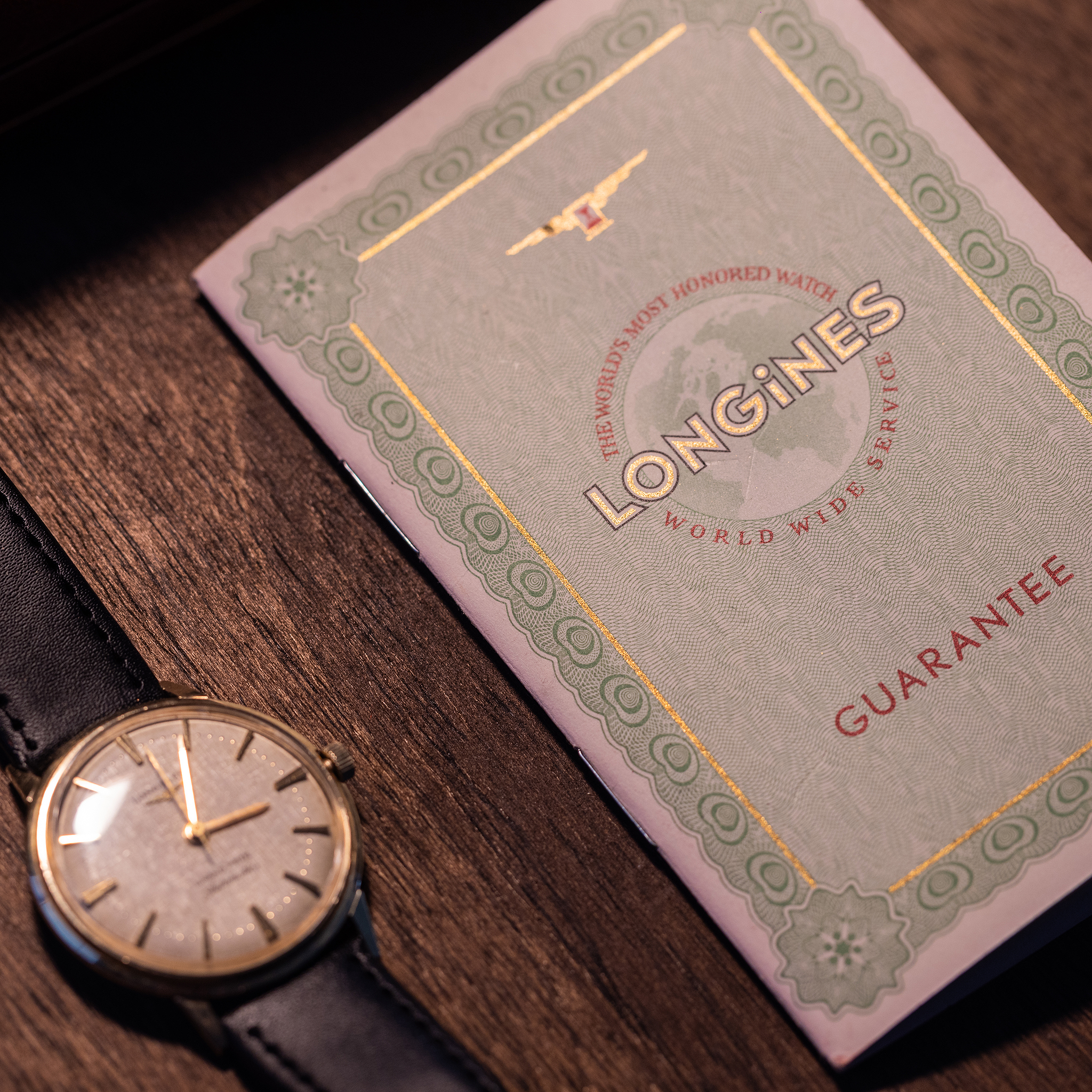 A Longines Grand Prize wristwatch with a black leather strap next to its guarantee booklet on a wooden surface.