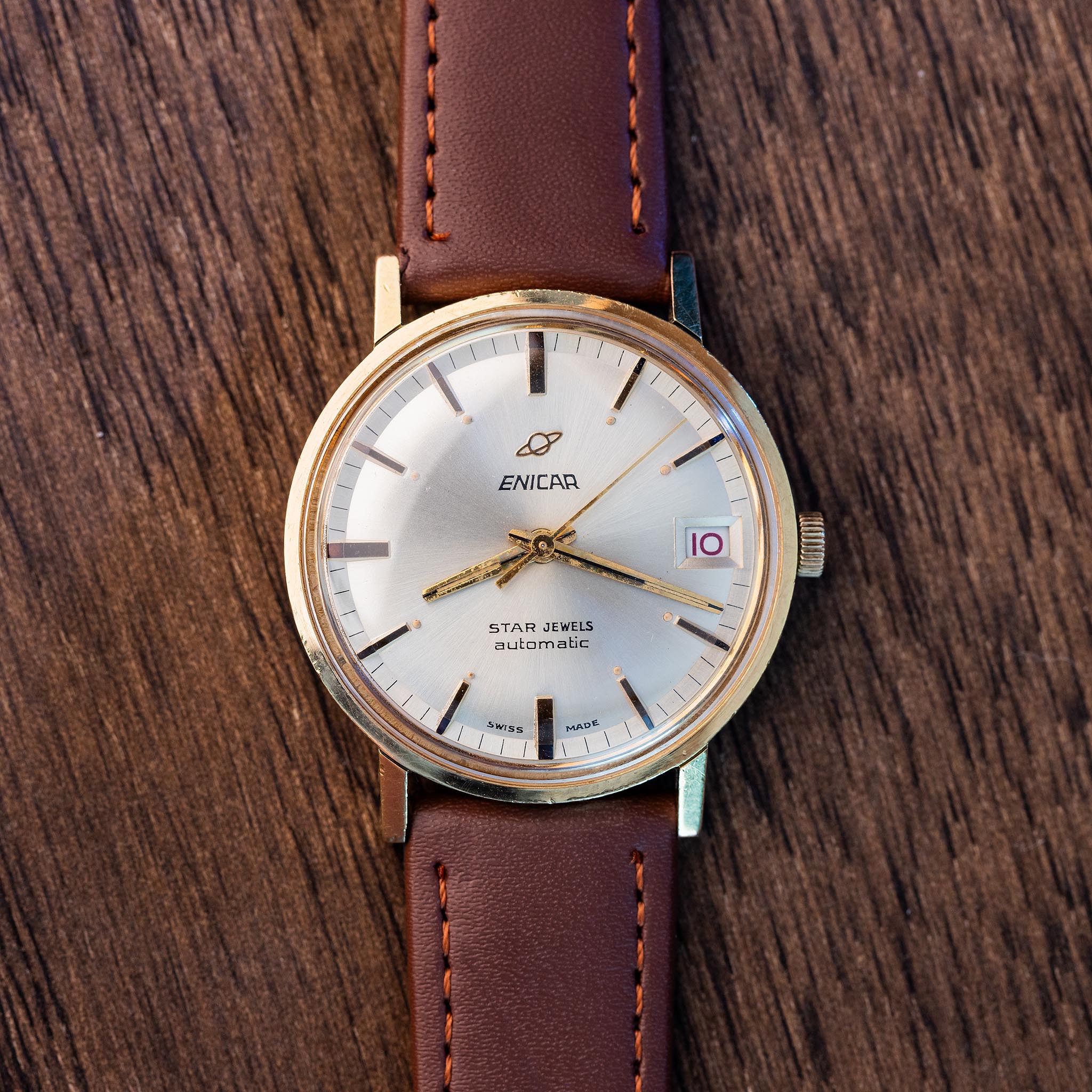 Vintage Enicar Ocean Pearl automatic wristwatch with silver dial, gold-tone case, red date numerals, and brown stitched strap photographed on wood.