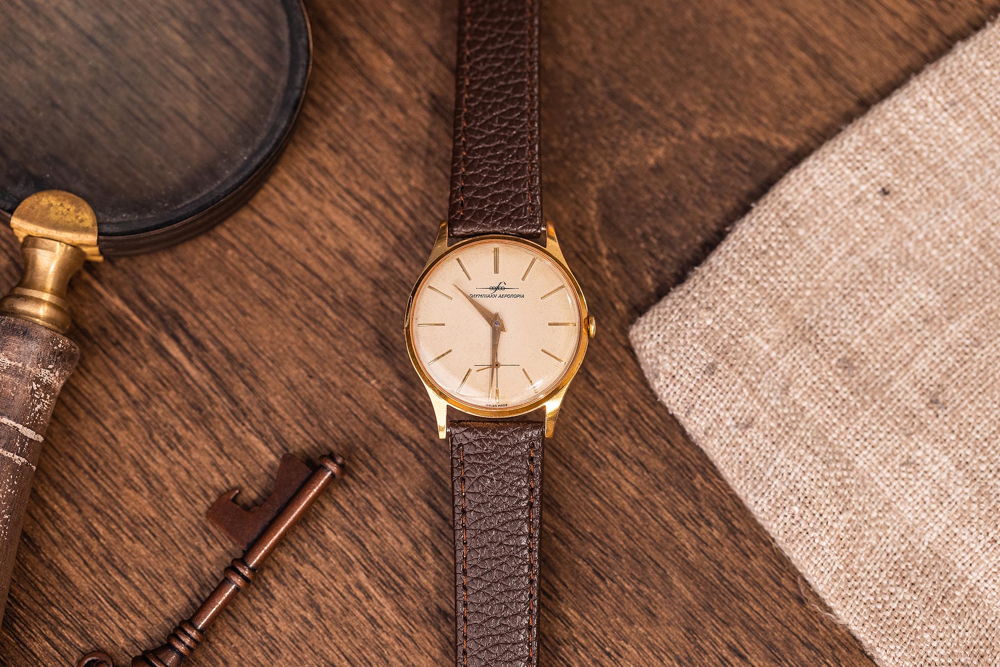 Full length vintage Breitling Olympic Airways Greek dial watch on brown strap arranged on wood with notebook and watchmaking tool.