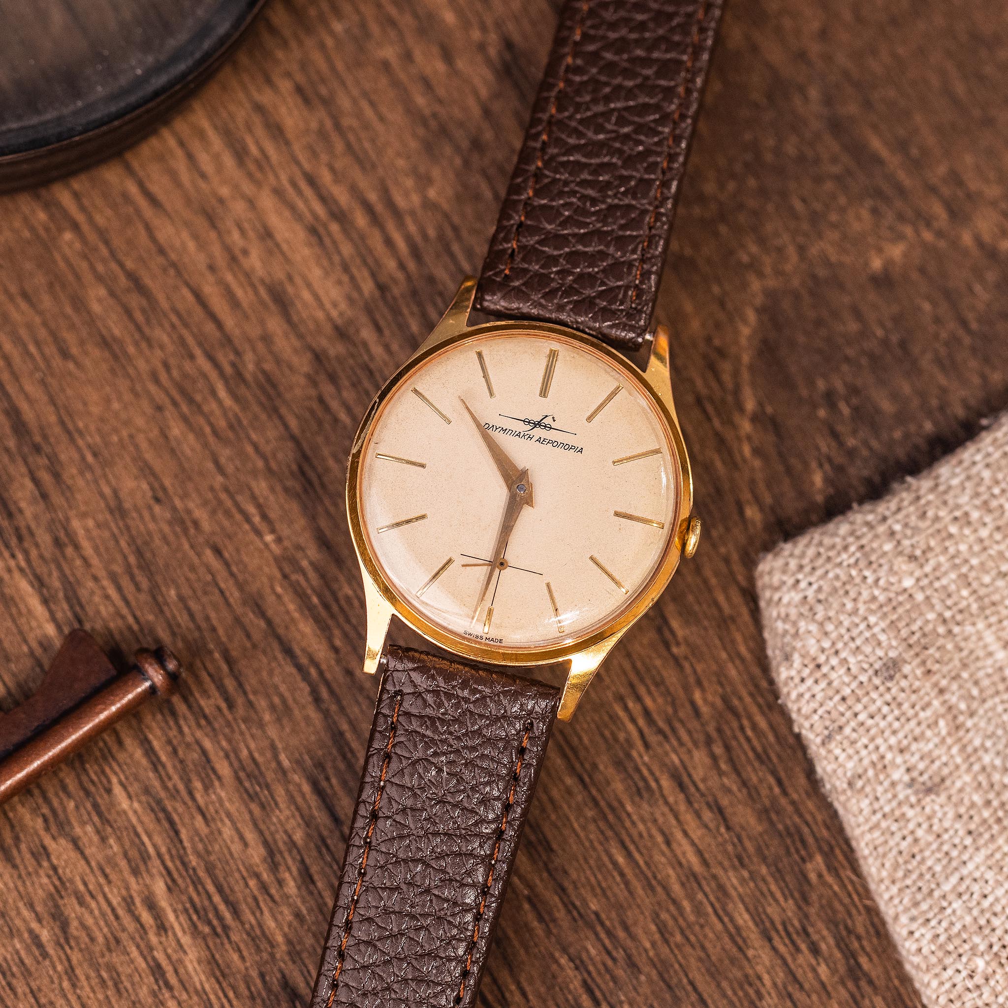 Gold-tone vintage watch on a brown leather strap, featuring a cream dial with the Olympic Airways logo and text.